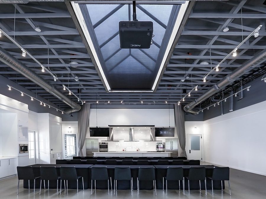The interior of Gainey Hall at Serenbe, its open room set with seating beneath a steel-truss ceiling and demonstration kitchen beyond