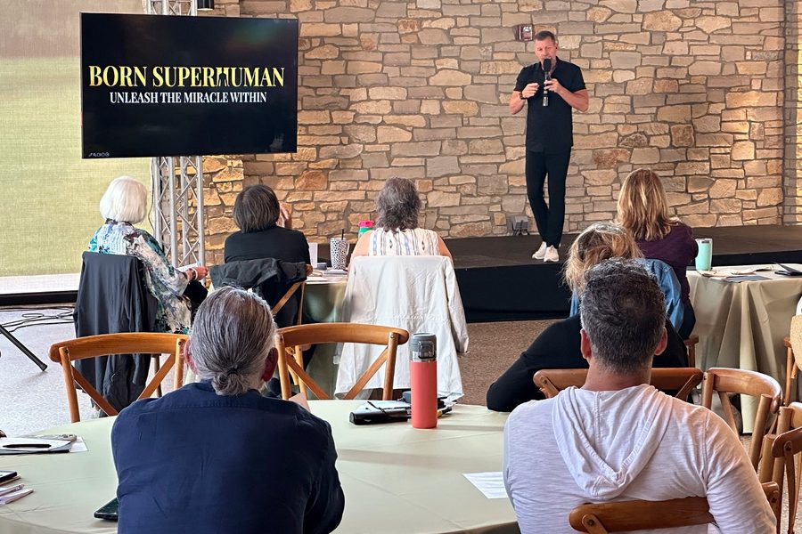 Dan Metcalfe on stage at the inaugural ALIGN Summit in June 2026, the BORN SUPERHUMAN title behind him and guests seated at round tables